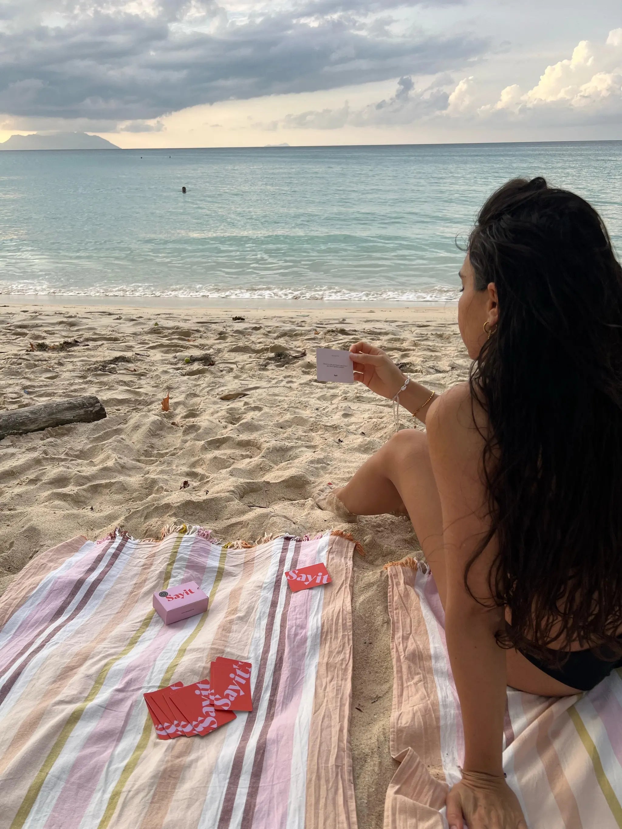 Frau am Strand hält eine SAYIT-Fragekarte, umgeben von Sand, Meer und Sonnenuntergang.